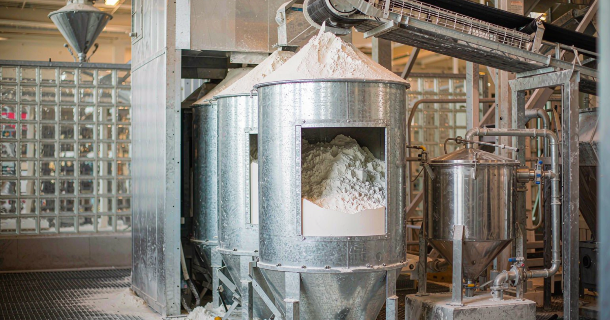 A glass factory batch house with storage silos of silica sand and soda ash feeding a weigh hopper before melting