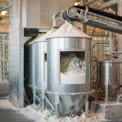 A glass factory batch house with storage silos of silica sand and soda ash feeding a weigh hopper before melting