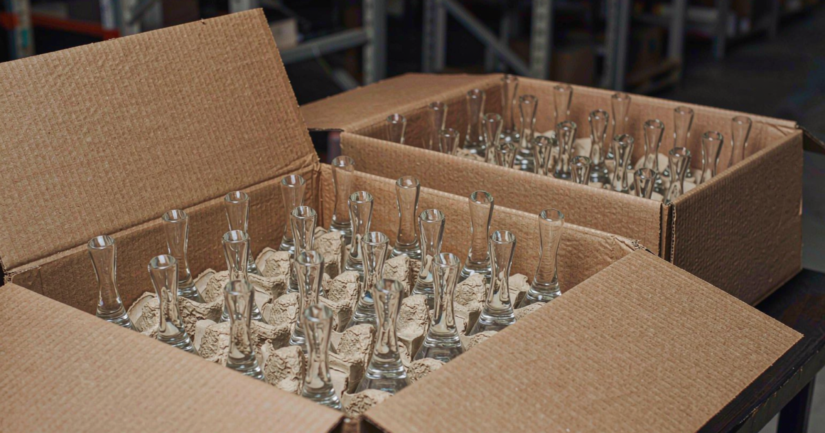Open corrugated shipping carton with molded pulp dividers holding rows of clear glass perfume bottles in a warehouse