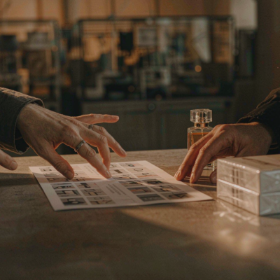 Two hands across a factory showroom table discussing a printed specification sheet beside clear glass perfume bottle samples