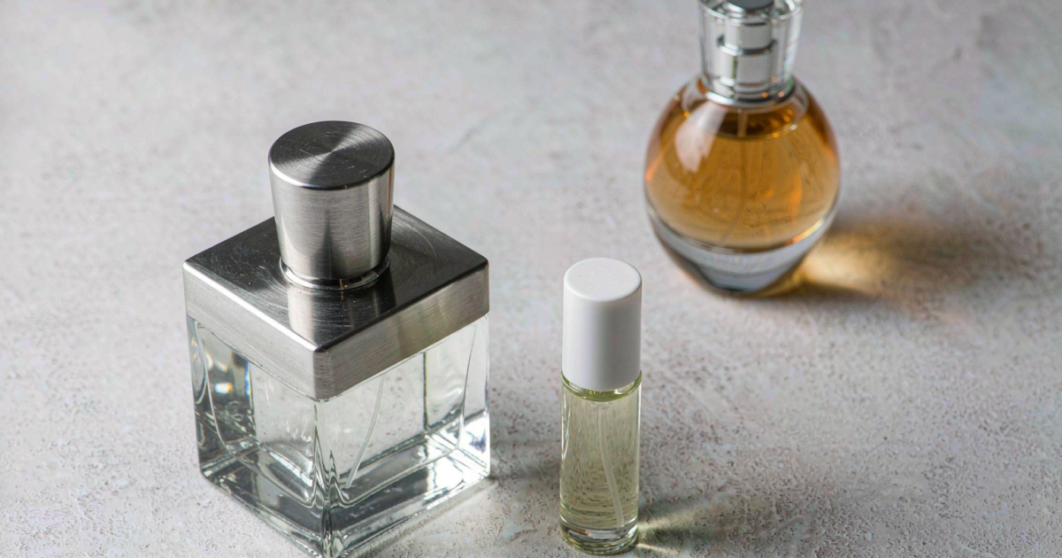 Three minimalist indie perfume bottles of different sizes on a light concrete surface, including a heavy square flacon with brushed metal cap and a small travel bottle