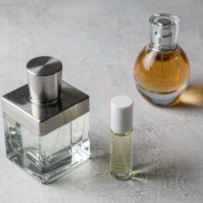 Three minimalist indie perfume bottles of different sizes on a light concrete surface, including a heavy square flacon with brushed metal cap and a small travel bottle