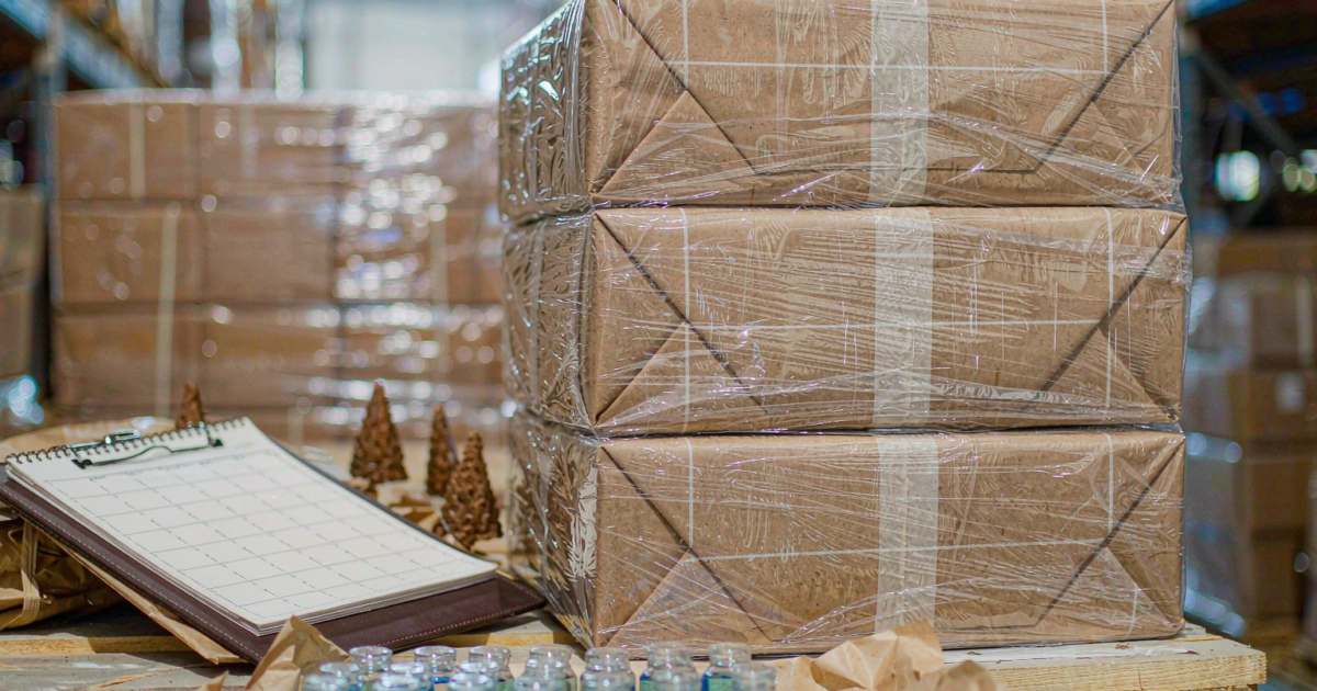 Stacked kraft cartons of glass perfume bottles on a wooden pallet with a clipboard and planning pad, clear flacons arranged in the foreground