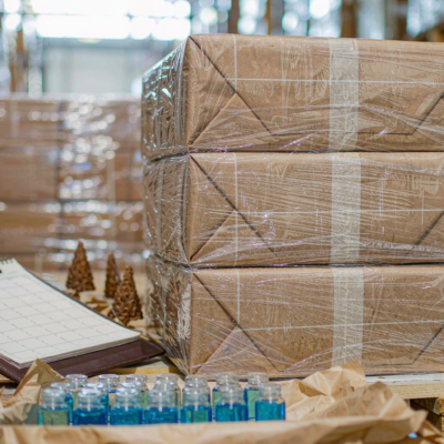 Stacked kraft cartons of glass perfume bottles on a wooden pallet with a clipboard and planning pad, clear flacons arranged in the foreground
