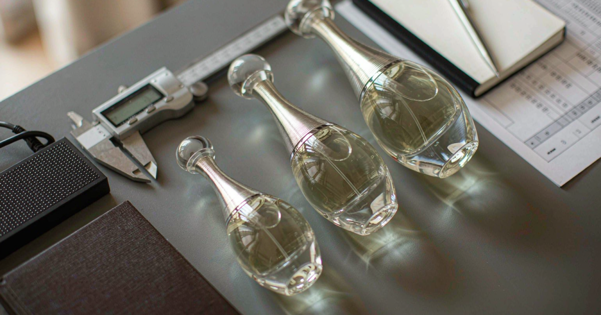 Three clear glass perfume bottles on a procurement desk beside printed quotation sheets and a digital caliper